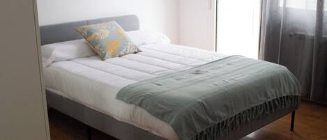 Un lit bien fait, avec une tête de lit grise, une couette blanche et un plaid vert.