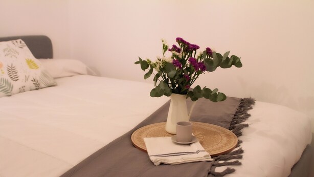 A neatly made bed with a white and gray bedspread, a decorative pillow, a vase with purple flowers, a small cup, and a woven tray.