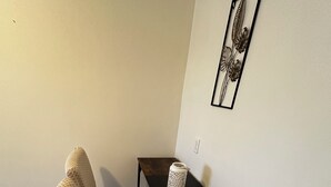 Ein kleiner, dunkler Holztisch mit einem weißen, gewobenen Stuhl. Auf dem Tisch steht eine weiße Vase mit gerolltem Rand. Ein gerahmtes Kunstwerk hängt an der Wand.