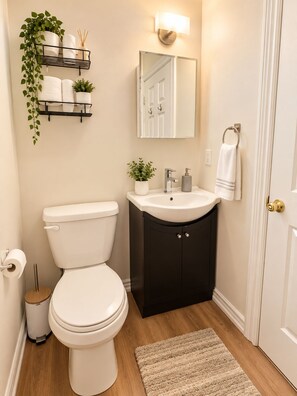 Baño con inodoro, lavamanos y espejo. Se observa una estantería con plantas y artículos de tocador.