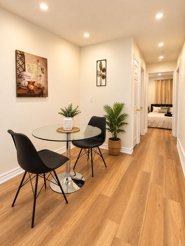A modern dining area with a glass table and black chairs, a painting on the wall, and a hallway leading to a bedroom.