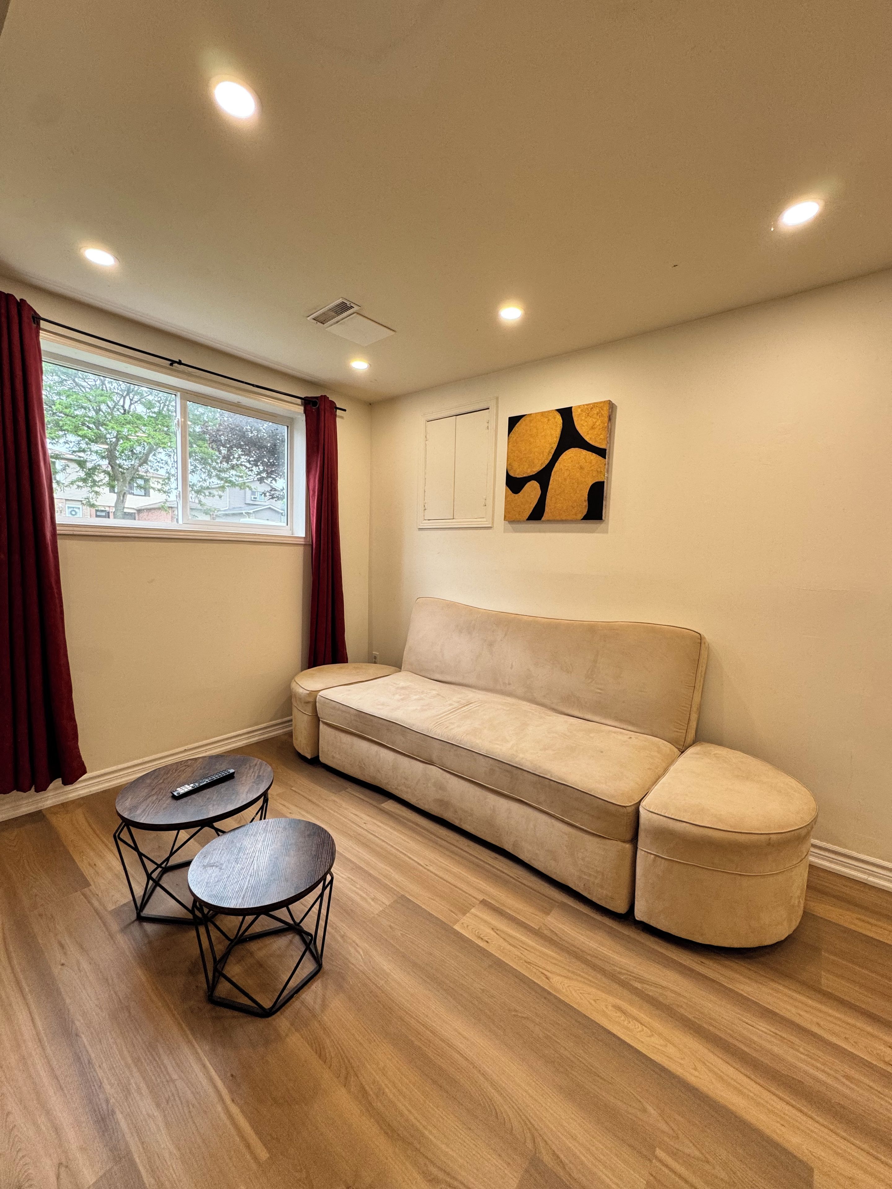 A room with a beige sofa, two round tables, a window with curtains, and a wall-mounted artwork.