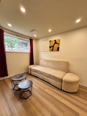 Una habitación con un sofá color beige, dos mesas redondas, una ventana con cortinas y una obra de arte colgada en la pared.