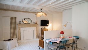 A room with a white wooden floor, a checkered wall hanging, a fireplace, a television, a dining table with chairs, and a ceiling fan.