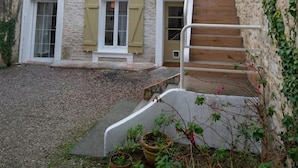 A two-story stone house with a wooden staircase, a small garden area, and a gravel driveway.