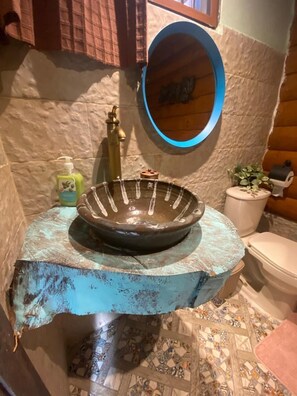 A bathroom with a rustic stone wall, a blue sink with a glass basin, a round mirror, a toilet, and a small potted plant. - Casa de Montaña Villa Maya (Jarabacoa)