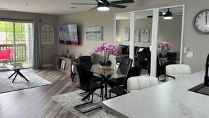 A modern living room with a dining area, featuring a large flat-screen TV, a ceiling fan, and a large clock on the wall. - BEAUTIFUL FULLY REMODELED WALK-IN 1 BEDROOM CONDO WITH FULL KITCHEN (Branson)