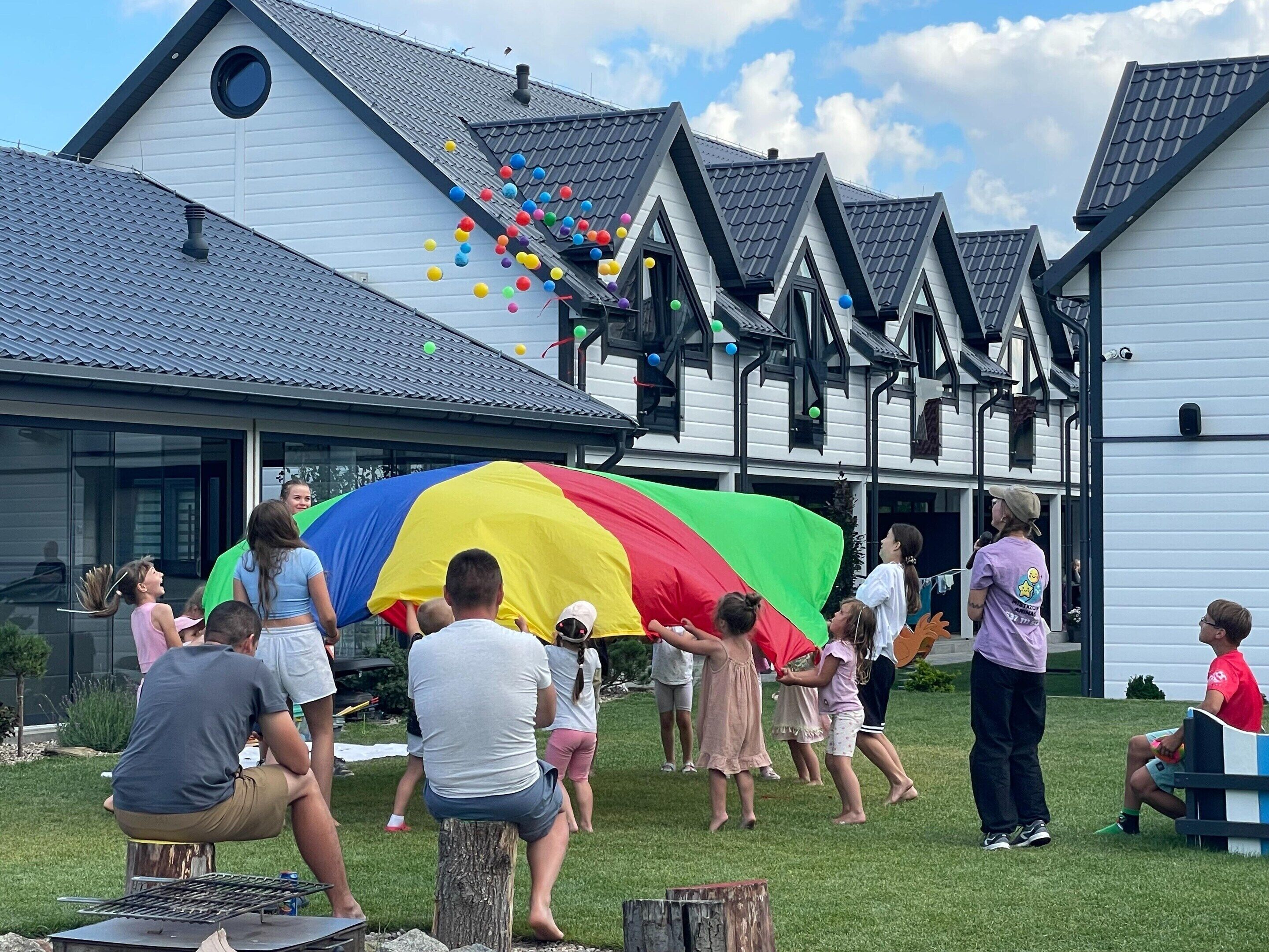 Een groep mensen staat verzameld rond een grote, kleurrijke parasol in een grasveld voor een gebouw met een puntige top en meerdere ramen.