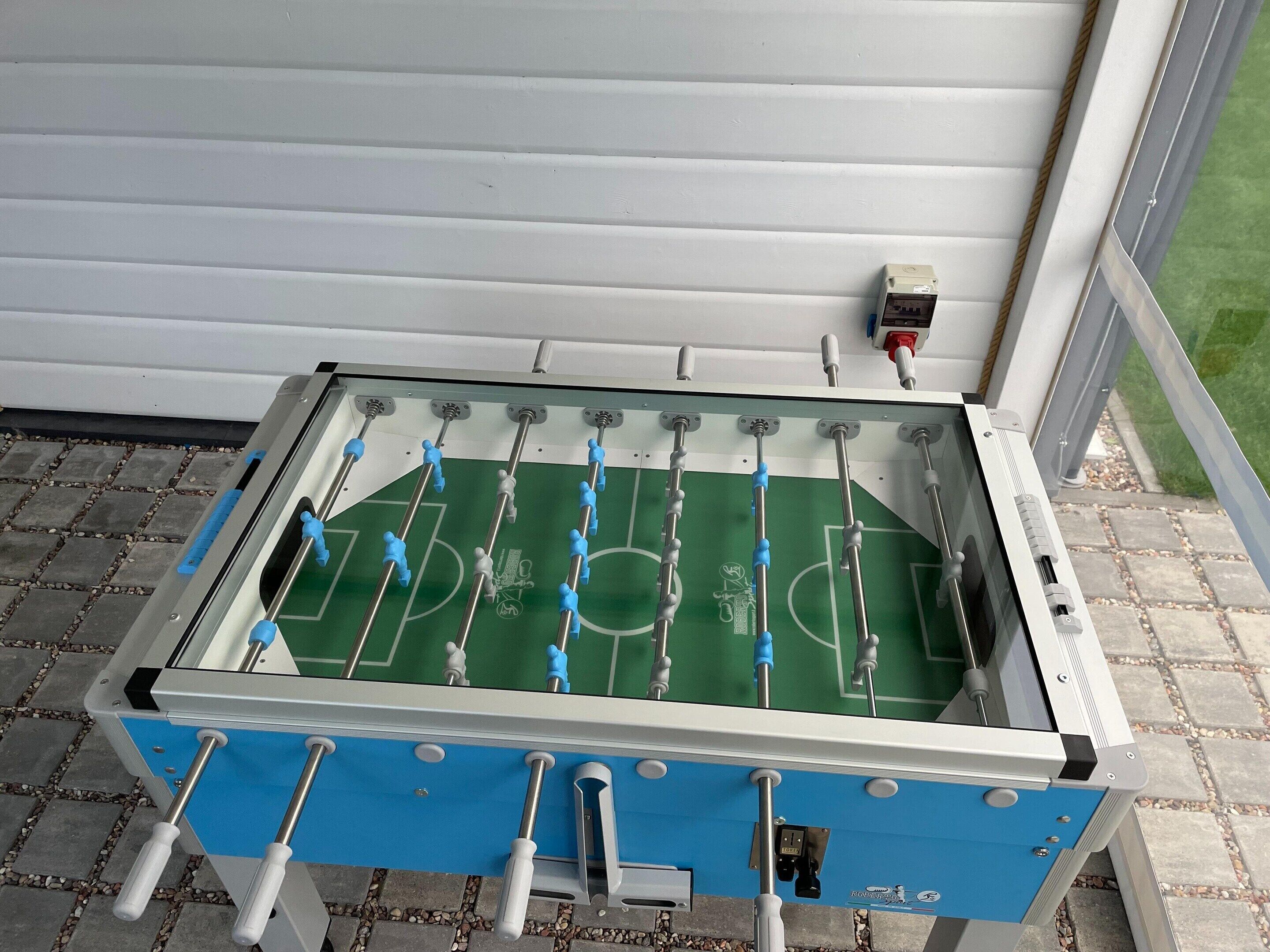A foosball table with a blue and white frame, positioned on a paved outdoor area with a white wall in the background.