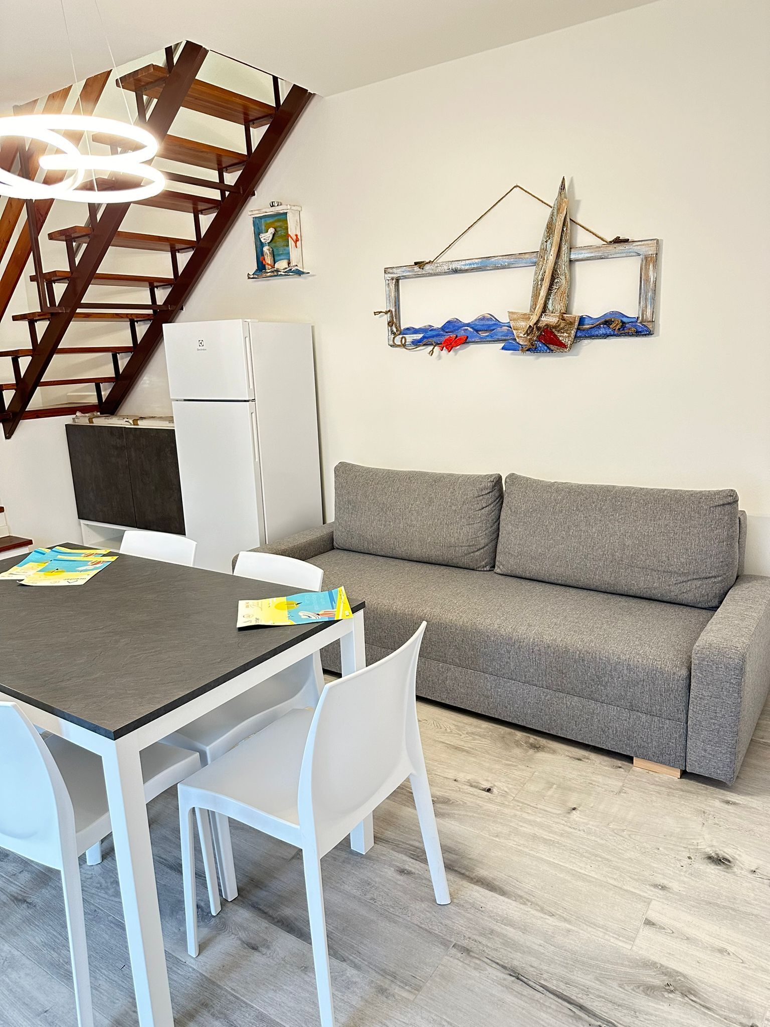 A modern living room with a grey sofa, white chairs, a wooden staircase, and a wall-mounted sailboat decoration.