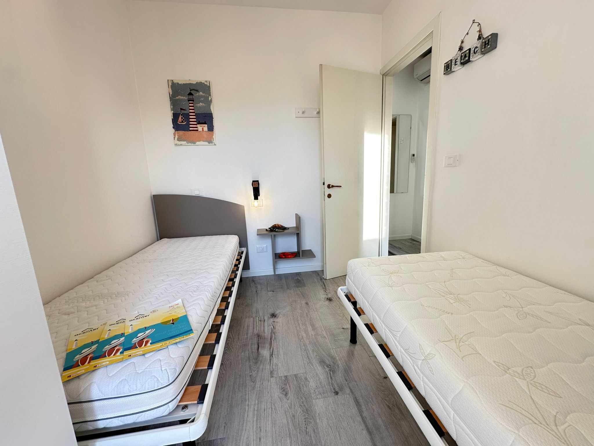A small, narrow room with two single beds, a nightstand, a wall-mounted lamp, and a door leading to another room.