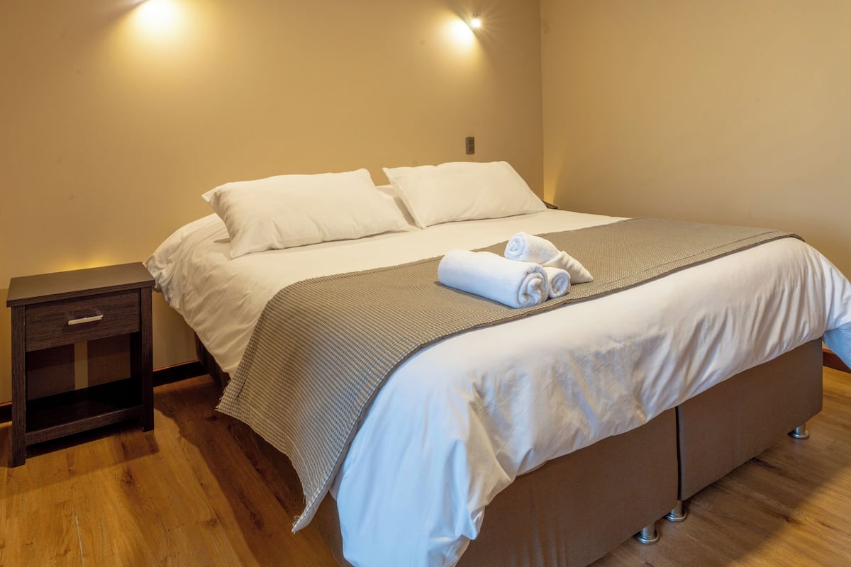 A neatly made bed with white linens and a brown bedspread, a small bedside table with a lamp, and an air conditioning unit on the wall.