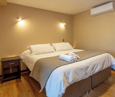 Una cama bien hecha con sábanas blancas y una colcha marrón, una mesita de noche con una lámpara y un equipo de aire acondicionado en la pared.