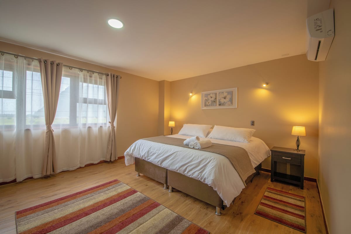 A bedroom with a bed, bedside lamps, a nightstand, a window with curtains, and a wall-mounted air conditioner.