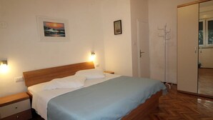 A bedroom with a wooden bed, white bedding, a nightstand, a wall-mounted light, a framed picture, a coat rack, and a wardrobe.