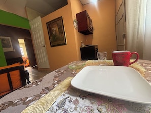 A room with a bed, a table with a white plate and a red mug, a framed picture on the wall, and a door leading to another room.