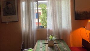 A room with a bed, a window with sheer curtains, a potted plant, and two framed pictures on the walls.