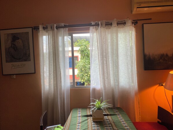 A room with a bed, a window with sheer curtains, a potted plant, and two framed pictures on the walls.