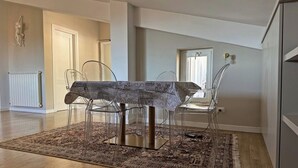 A dining area with a table covered by a patterned tablecloth, surrounded by clear chairs. A large, patterned rug covers the floor, and a radiator is visible in the background. - Attic with terrace and nice view! (Sora)