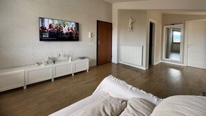 A modern living room with a flat-screen TV mounted on a white brick wall, a white console table with decorative items, wooden flooring, and a large window offering a view of the outdoors. - Attic with terrace and nice view! (Sora)