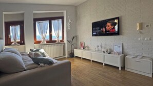 A modern living room with a large flat-screen TV mounted on a white brick wall, a sofa with cushions, a white console table, and a view of the sea through large windows with curtains. - Attic with terrace and nice view! (Sora)