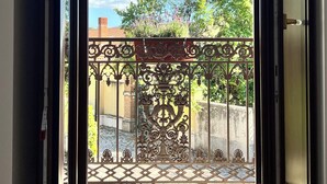 Une porte-fenêtre coulissante s’ouvre sur un balcon agrémenté d’une rambarde en métal décorative, d’où l’on peut admirer des arbres et un bâtiment en arrière-plan.