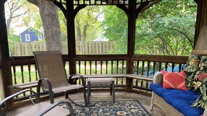 A covered outdoor seating area with a wooden bench and chairs, a patterned rug, and a view of a blue house through the wooden railing.