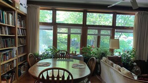 A dining area with a round table, chairs, a bookshelf, and large windows with a view of greenery.