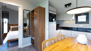 A compact hotel room with a bed, a wooden wardrobe, a dining table with chairs, a kitchen area with a stove, and a large window. - Waterfront Retreat, Friesland (Delfstrahuizen)