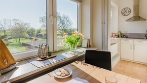 Una cocina con vistas a un parque, una mesa de comedor con un plato de pasteles, un vaso de zumo de naranja y un jarrón con flores.