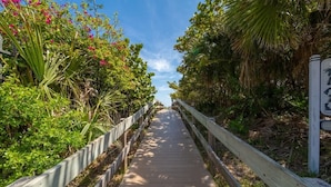 A wooden boardwalk flanked by lush greenery and flowering plants, leading towards a building. - Oceanfront I Resort-Style I Heated Pool I Beach (Cape Canaveral)