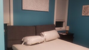 Un dormitorio con cama, dos almohadas, una mesita de noche, una lámpara y una obra de arte colgada en la pared.