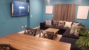 Una sala de estar con un televisor montado en la pared, un sofá con cojines estampados, una mesa de comedor con sillas y una planta.