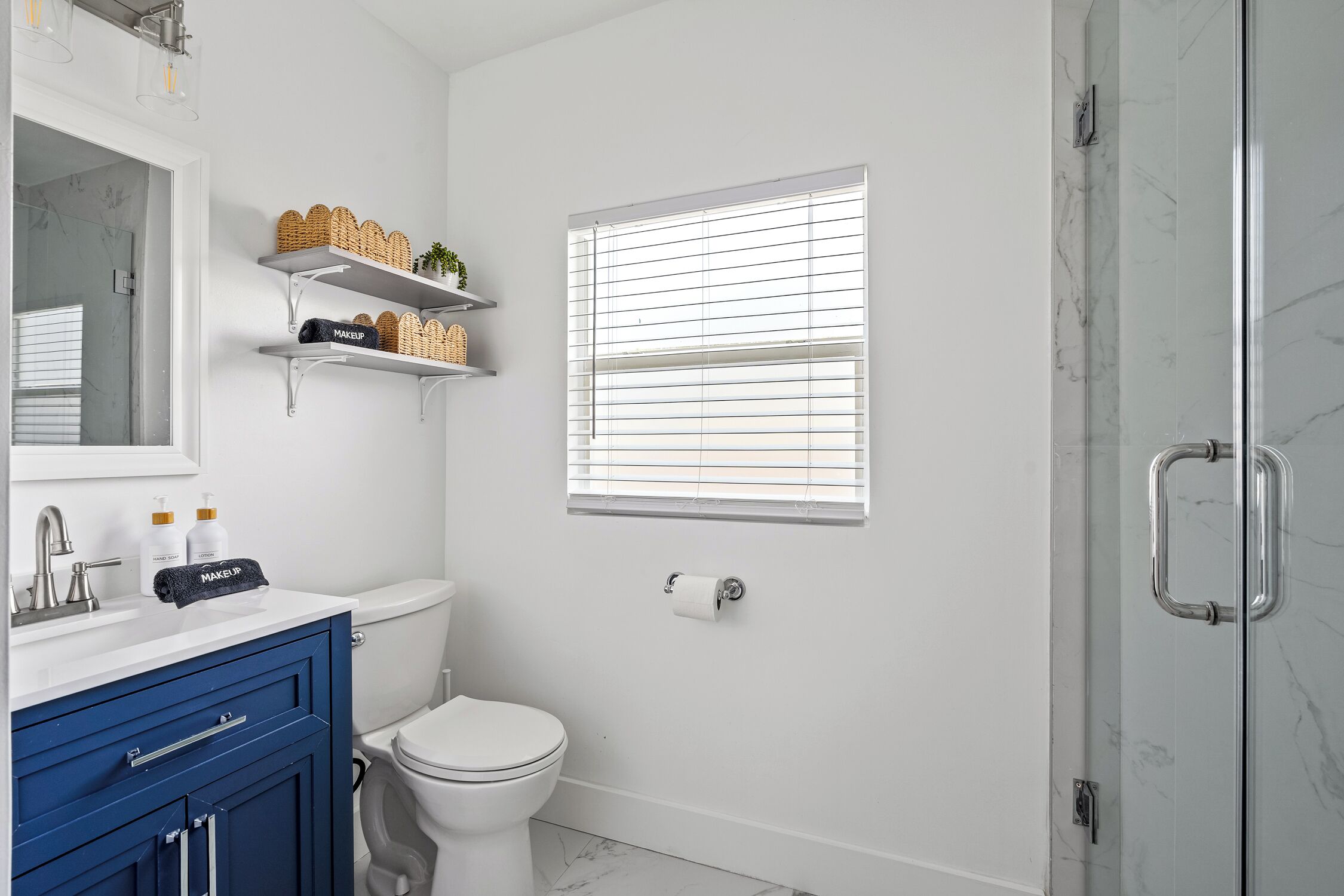 A bathroom with a white toilet, a blue vanity, a mirror, and a window with blinds.