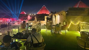A rooftop bar with tables and chairs, a view of the pyramids, and a stage with a crowd.