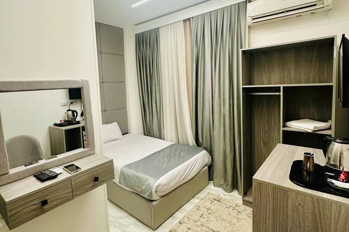 A modern hotel room with a bed, a desk, a chair, a mirror, and an air conditioning unit.