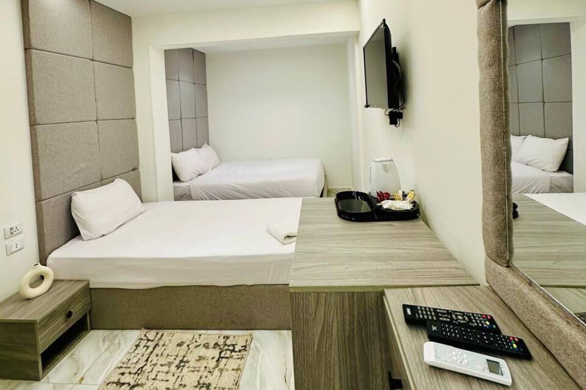A modern hotel room with a large bed, a desk with a computer, a mirror, and a small side table.