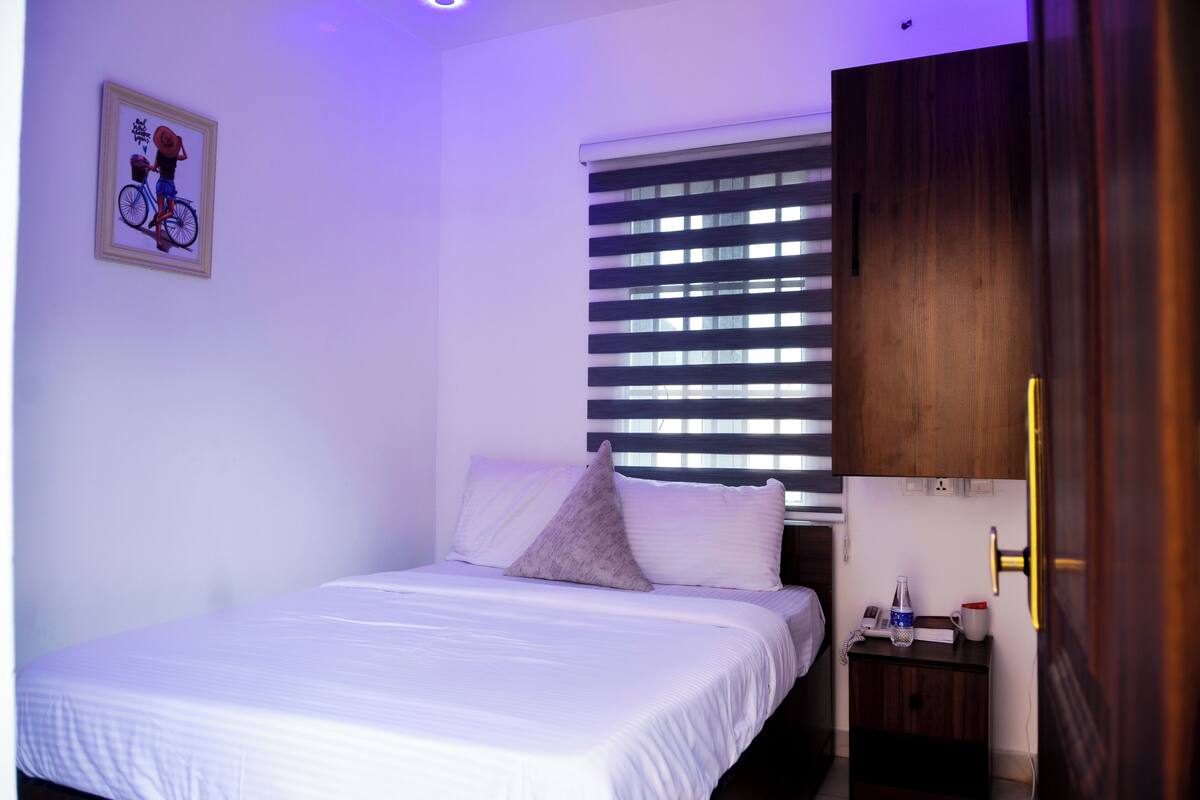 A neatly made bed with white linens, a small bedside table with a lamp, a framed picture on the wall, and a window with blinds.