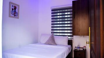 Cama bem arrumada com lençóis brancos, uma pequena mesa de cabeceira com uma luminária, um quadro na parede e uma janela com persianas.