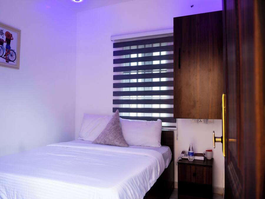 Cama bem arrumada com lençóis brancos, uma pequena mesa de cabeceira com uma luminária, um quadro na parede e uma janela com persianas.