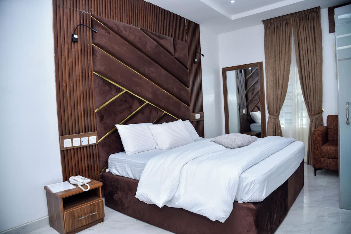 A modern bedroom with a large bed, a wooden headboard, a bedside table with a phone, and a mirror.