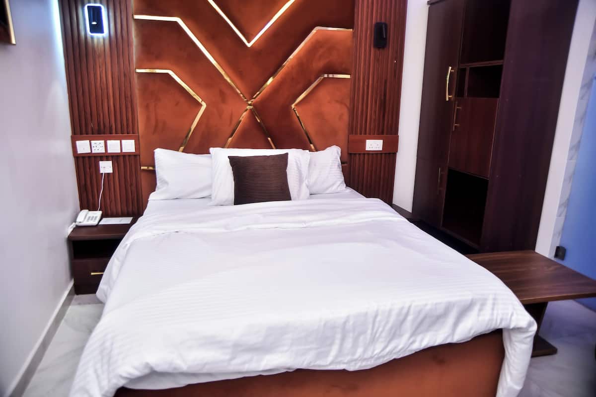 A neatly made bed with white linens and a brown accent pillow, a wooden headboard with geometric light fixtures, and a bedside table with a phone and a notepad.