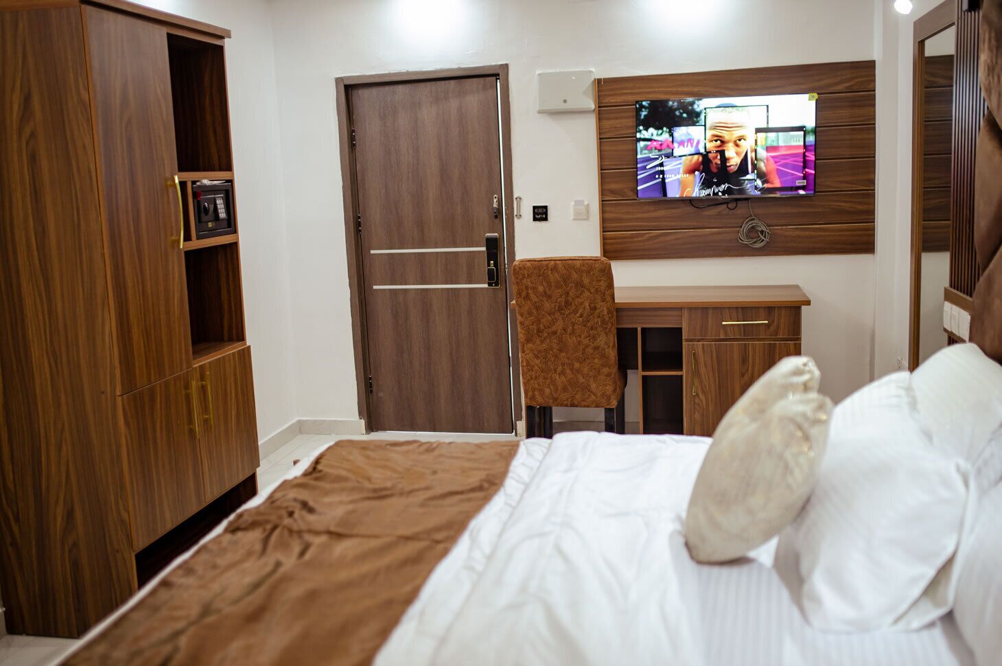 A hotel room with a bed, a desk, a chair, a TV mounted on the wall, and a wardrobe.