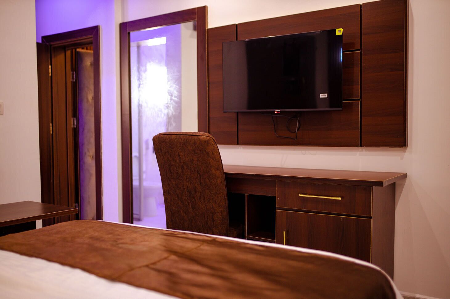 A hotel room with a bed, a desk, a chair, a TV mounted on the wall, and a door with a purple light.