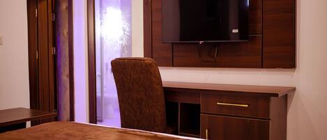 A hotel room with a bed, a desk, a chair, a TV mounted on the wall, and a door with a purple light.