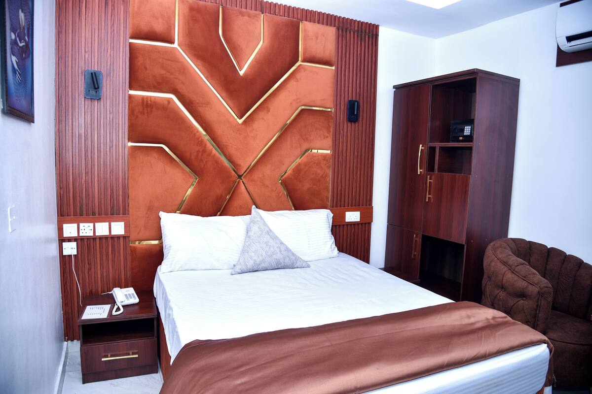 A neatly arranged hotel room with a large bed, a wooden headboard, a wardrobe, and a bedside table with a phone.