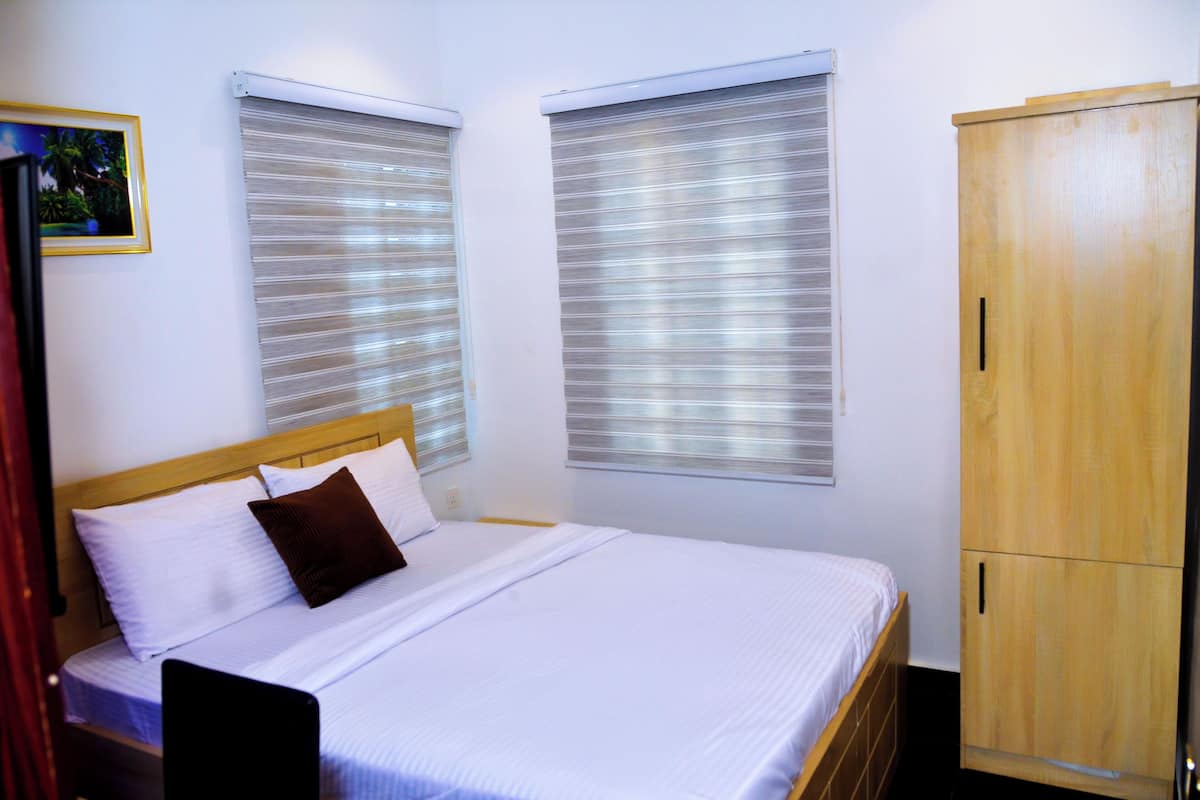 A bedroom with a bed, two windows with blinds, a wooden wardrobe, and a framed picture on the wall.