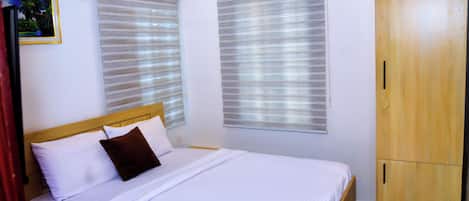 A bedroom with a bed, two windows with blinds, a wooden wardrobe, and a framed picture on the wall.