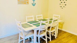 Un comedor con una mesa y sillas blancas, un ancla decorativa en la pared y una cita enmarcada.