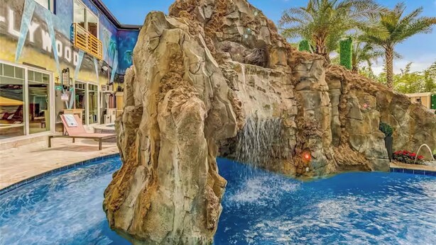 Une piscine agrémentée d’une cascade artificielle en roches, de palmiers et d’un bâtiment sur lequel on peut lire l’inscription « Wood ».